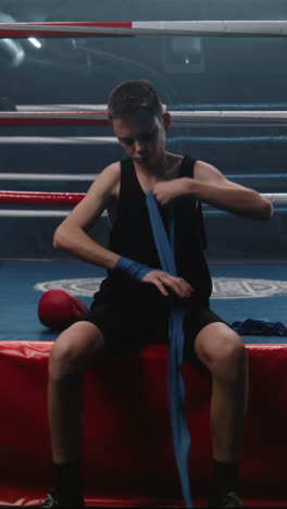 Young-boxer-sits-near-boxing-ring-and-wraps-his-hands-with-bandage-for-martial-arts.-Teenage-athlete-prepares-to-fight-tournament-or-competition,-trains-in-boxing-gym.-Physical-activity.-Vertical-shot
